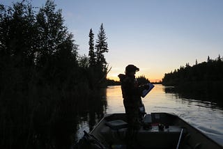 A person stands in a boat in silhouette with forest and water and the beginning of a sunrise in the background.