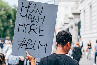A Black man at a protest faces away from the camera holding a hand-written sign that says “How Many More #BLM”