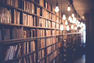 A stack of bookshelves on the left, illuminated by lightbulbs, with light streaming in from a window in the far distance.