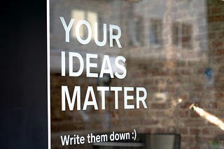 White letters on a window. The letters read “YOUR IDEAS MATTER” and then “Write them down :)”. The reflection of stone buildings can be seen on the window.