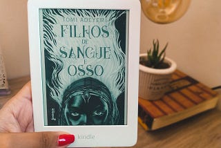 Foto de uma mão segurando um aparelho Kindle na cor branca, exibindo a capa do livro filhos de sangue e osso. Ao fundo aparece, desfocado, um livro com um jarro bege com uma suculenta e acima, aparece parte de uma lâmpada.