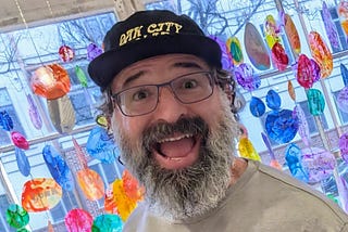 Closeup of the author with a big open-mouth smile. He’s wearing a hat, has a long graying beard and glasses, and is standing in front of colorful glass art.