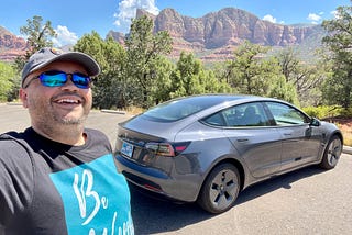 The author taking a selfie with the Tesla Model 3 in the background and beautiful Sedona mountains in the distance.