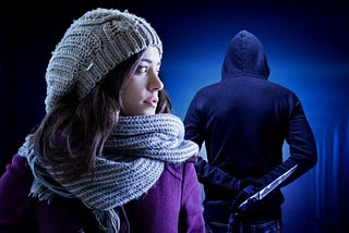 A girl in winter clothing looks nervously over her shoulder at a shadowy stalker in the background. He is facing away and holding a knife.