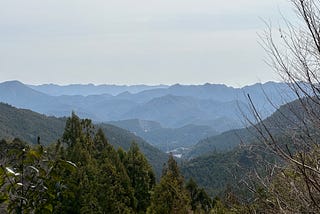 A viewpoint looking down on one of the Kumano Grand Shrines. It is taken from a height, and you can see the mountain range in the background and the valley with the shrine in the front.