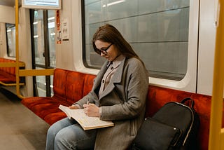 woman in grey coat with black backpack sitting on train writing in journal