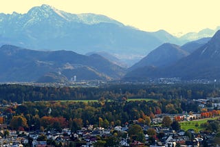A residential area is in the foreground, surrounded by alpine trees in a valley surrounded by huge, snow-capped mountains.