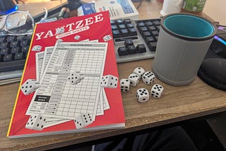 An off-brand Yahtzee scorepad (the “H” is covered by a rolling die, to avoid trademark offense), six dice and a leatherette dice cup sitting on a desk in front of a computer keyboard.