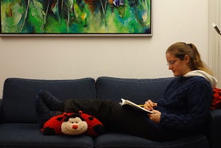 Erlijn van Genuchten sitting on a sofa, reading a book. She is wearing soft boots, comfortable ski trousers and two pullovers to stay warm in a chilly house.
