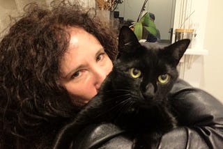 A woman with long, curly black hair wearing a black leather jacket (Karen Dendy Smith) and holding a black cat with yellow eyes (Hockey Puck).