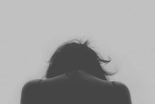 A black and white photograph of a sad woman facing away from the camera. Her head is lowered.