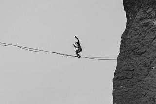 Looking up from below to see a man in the distance walking on a tightrope from the edge of a cliff.