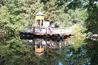 A picture of a boat dock with green trees surrounding it and a small wooden tower on the dock and tires on the front. The water shows a complete detailed reflection of the image. It is hard to tell which way to hold the photo, it is so clear.