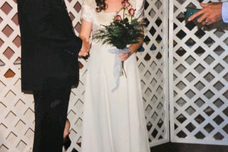 The image is an author provided picture of her wedding as an 18 year old woman to a 30 year old man. Her expression is unreadable, but not the happy expression you’d expect of a bride. His head is out of the frame. She is holding a bouquet of roses and the officators hands and bible are visible to the right side of the frame. A closer look reveals the bride’s child hiding behind the groom’s legs.