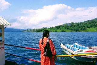 Woman gazing at a boat in the water with mountains and greenery in the distance