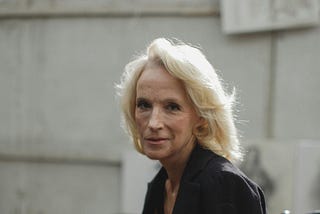 A woman with white hair and wrinkles, looking directly at the camera with a pensive look on her face, against a cement block wall.