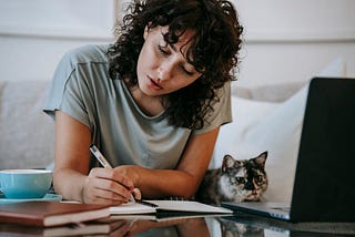 A young woman with dark curly hair writes in a notebook. Next to her, a gray cat looks at a laptop.