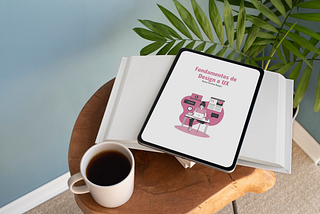 Uma mesa de canto com um livro, um e-reader e uma xícara de café. No e-reader está a capa do ebook escrito “Fundamentos de Design e UX” e o nome da autora “Bruna Cristina Berges”. Ao fundo da imagem tem uma planta no ambiente.