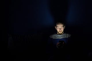Main sits in darkness, light of phone illuminates his face.