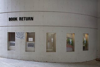 An exterior curving light tan concrete structure with a “Book Return” sign at the left side. Underneath the sign, there are two book returns. To the the right, there are three small modern glass panels offering a peek into the library.