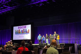 A dark purple stage, a slide that says SIGCSE, and the organizing committee kicking things off.