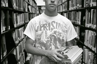 Young man wearing Bob Marley Uprising shirt stands in a library holding books.