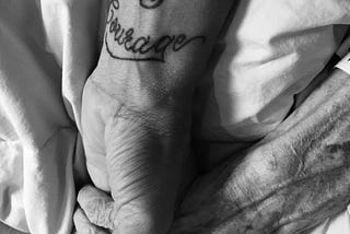 A young hand with “Strength and Courage”   tattooed on the inner wrist holds an older hand.