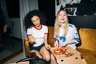 Two women eating pizza and drinking wine.