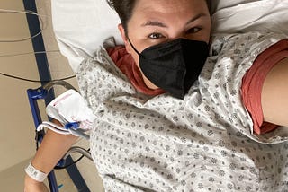 A white woman in her thirties takes a selfie of herself in a hospital bed. She is wearing a hospital gown over her shirt and a mask on her face, and she is hooked up to lots of tubes