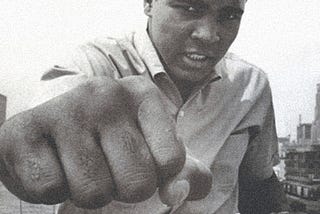 Boxer, Muhammad Ali, throws a punch, looking intently at the camera, with his teeth clenched.