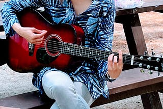 A red guitar. The woman playing the guitar is sitting on a picnic bench and her face is not in the frame