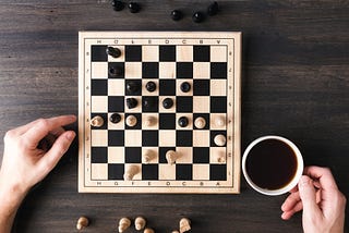 A chess game on a small board framed by two hands and a cup of coffee.