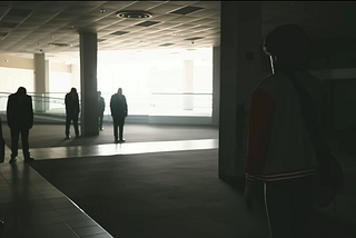 A screenshot from Generation Loss, after the button press. A wide shot, in a dimly lit room, with ambient light from outside. Around the room are several employees, now no longer moving. Ranboo begins walking to the left. In the corner, a small lit exit sign, pointing to the right.
