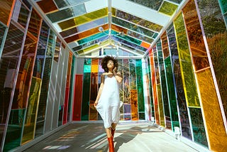 A woman stands smiling in a multicolored, stained glass atrium wearing a white dress and red boots.