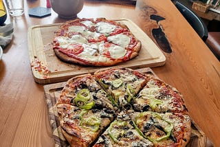 Two homemade pizzas on a wooden table with a bottle of wine in the background