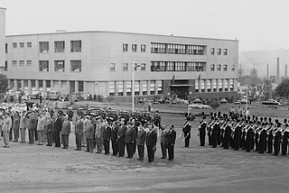 Black-and-white photograph of officials standing in lines outside buildings.