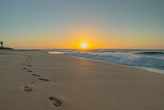 A landscape featuring beach and surf and a perfectly clear sky. A line of footprints in the sand stretches backwards from the viewer along the beach towards the setting sun