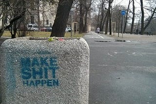 Words “Make Shit Happen” on concrete planter in front of tree on walkway.