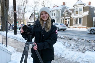 Lauren Weseluck holds a camera on a tripod on the sidewalk with the street and older-style houses in the background.