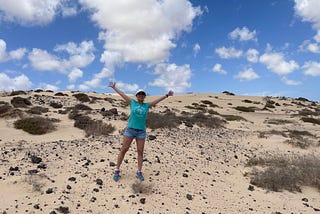 A lady in a teal shirt and denim shorts with a white cap having her hands stretched out like she’s doing a star jump. The backdrop is a sand dune with specks of dessert weeds and pebbles all over, shadowed by blue skies with spots of cotton white clouds.