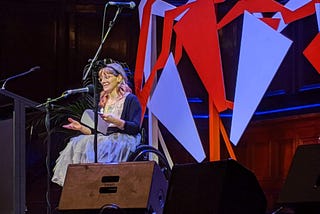 image of a woman in a wheelchair on stage reading, she is wearing a tutu, glasses and has long pink curly hair