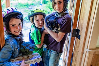 Three smiling children in bicycle helmets, standing in a doorway, one holding a cat and another holding a carton of eggs and broccoli.