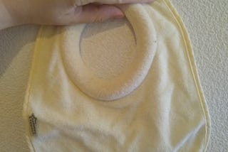 A hand holds a Tommee Tippee brand bib up to a white plaster wall. The bib is a soft yellow colored cloth. The collar of the bib has a thick cushion around where baby’s neck would go, which is signature of this particular bib.