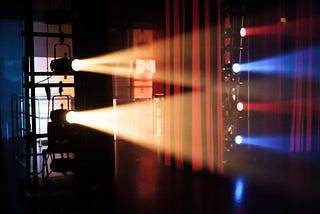 Backstage theater lights with a theater curtain in the background. To the left, two big yellow lights project horizontally towards the right. To the right, smaller alternated red and blue lights also project towards the right.