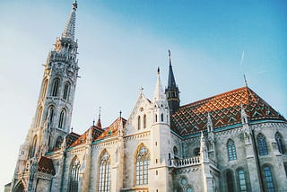 A gothic Hungarian cathedral is shown on a clear, sunny day. The walls are white and the roof is the color of clay. The cathedral has many towers and stained glass windows. This is what I imagine the setting of the Matula Academy in the book to look like.