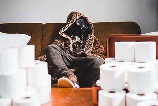 A person wearing a hooded jacket and camo pants sitting on brown couch with a gas mask, surrounded by stacks of toilet paper.
