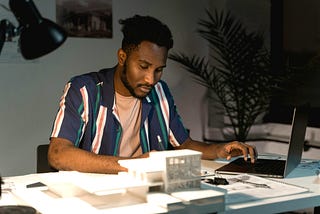 A man with short curly hair and a trimmed beard sits at a desk, focused on his work. He is wearing a navy shirt with vertical red, white, and green stripes over a beige T-shirt. In front of him are architectural models and papers, and he is using a laptop with one hand while holding a pen in the other.