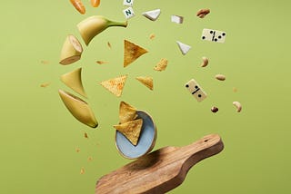 A banana chopped in random pieces tossed up in the air with dominoes, nachos and some scrabble alphabets along with it from a wooden chopping board.