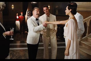 Kenneth Branagh (as Hercule Poirot) wearing a white tux and huge mustache takes Gal Gadot’s (Linnet Ridgeway)’s hand. She is wearing a white dress. Tom Bateman in a white suit is in the background between them, smiling.