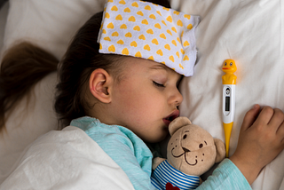 A young child is asleep in bed with a wet cloth on their forehead, a plush monkey toy in their arm, and a digital thermometer next to them.